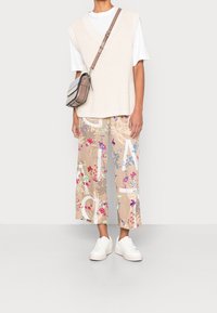 Beige gestrickte Weste über einem weißen T-Shirt, kombiniert mit weiten Hose mit floralem Muster in Beige, Pink und Blau. Weiße Sneaker und kleine Umhängetasche.