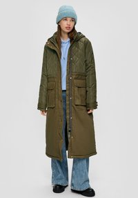 Lang olijfgroen gestept jasje met een capuchon, voorzien van zijzakken en een rits, gedragen over een lichtblauwe hoodie en denim broek.