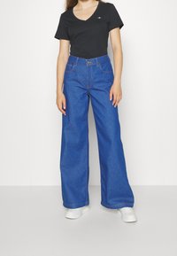 Modré džíny s širokými nohavicemi a vysokým pasem, s předními kapsami a kontrastním švem, v kombinaci s černým tričkem s výstřihem do V.