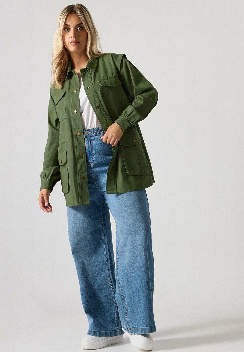 Vaaleahiuksinen nainen, joka on pukeutunut ylipitkään vihreään napitettuun takkiin, valkoiseen paitaan, leveisiin sinisiin farkkuihin ja valkoisiin tennareihin tasaisella taustalla.