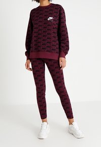 Burgundy Nike sweatshirt med djärvt svart logotyp och textmönster, i kombination med matchande leggings. Vita sneakers kompletterar looken.