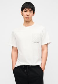 Λευκό βαμβακερό μπλουζάκι με υφασμένη γκρι τσέπη που έχει επαναλαμβανόμενο μοτίβο "MK". Μαύρα γράμματα εμφανίζουν το "MICHAEL KORS" στο μπλουζάκι.