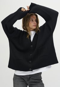 Čierny oversized cardigán s rebrovanými manžetami a lemom, s V-výstrihom, veľkými gombíkmi a prednými vreckami. Nosený cez biely top.