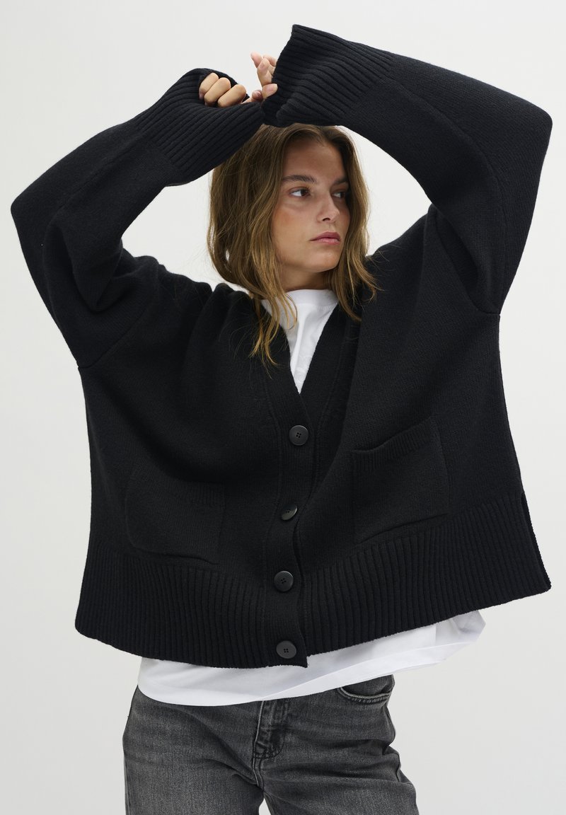 Čierny oversized cardigán s rebrovanými manžetami a lemom, s V-výstrihom, veľkými gombíkmi a prednými vreckami. Nosený cez biely top.