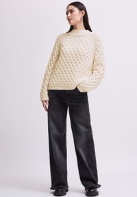 Jeune femme portant un pull en laine texturé crème, un jean noir à jambes larges, des chaussures noires épaisses et des boucles d'oreilles en argent, se tenant avec une main dans la poche.