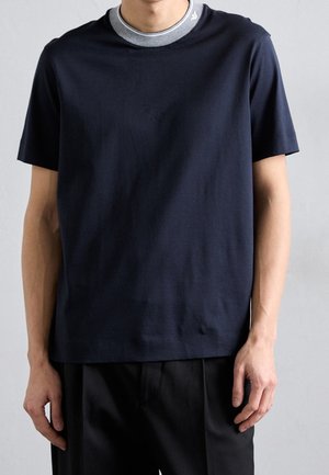 Mann trägt ein marineblaues T-Shirt mit kurzen Ärmeln über einem grauen Rundhalsausschnitt-Unterhemd, kombiniert mit schwarzen Hosen vor einem schlichten Hintergrund.