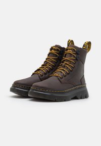 Botas de tobillo de cuero marrón con cordones amarillos, una suela negra gruesa y una etiqueta amarilla con el nombre de la marca. Presenta costuras detalladas.