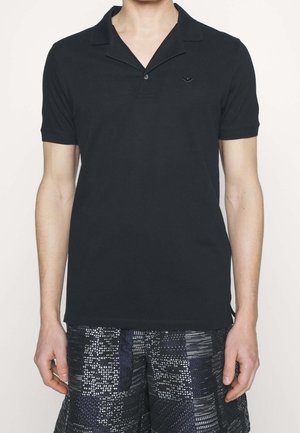 Mann trägt ein schwarzes Poloshirt mit kurzen Ärmeln und kleinem Emblem auf der Brust sowie gemusterte dunkle Shorts.