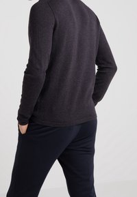 Maglione grigio scuro a maniche lunghe, tessuto morbido, design aderente; abbinato a pantaloni blu scuro, in piedi con le mani nelle tasche.