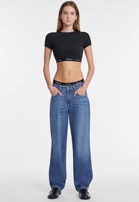 T-shirt noir court et jean bleu en denim à coupe ample. Le denim présente un lavage clair avec un léger effet délavé. Le mannequin se tient les mains dans les poches.