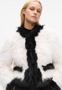 Faux fur jacket in white and black, featuring a shaggy texture, wide collar, and contrasting panels. Embroidered logo on the neckline.