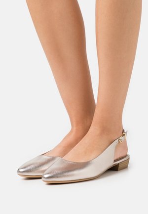 Des pieds portant des ballerines argentées métalliques avec des bouts pointus et de petits talons en bois sur un fond blanc.