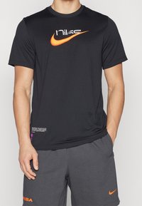 Muž oblečený v černém tričku Nike s oranžovým logem swoosh a tmavě šedých šortkách s oranžovými logy NBA a swoosh, stojící s jednou rukou v kapse.