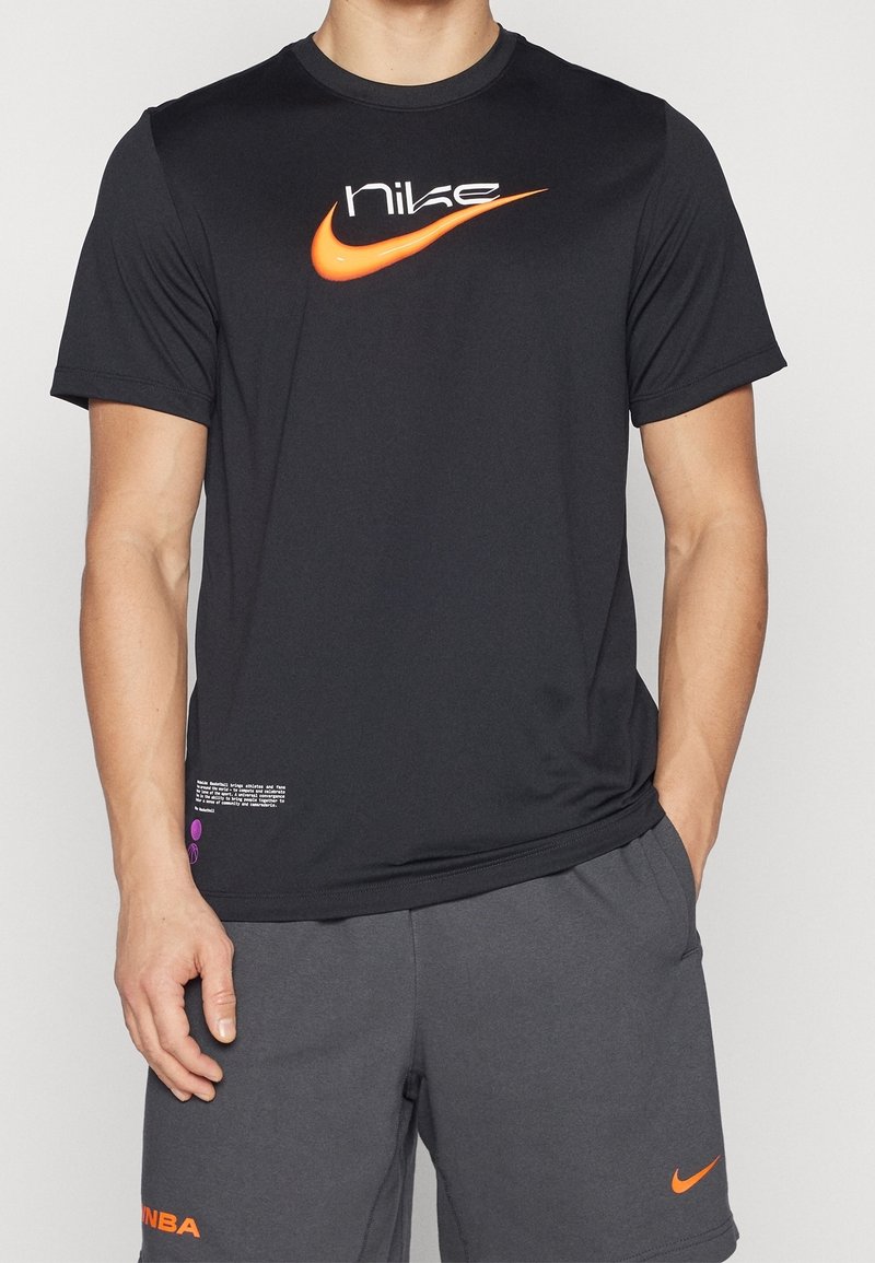 Muž oblečený v černém tričku Nike s oranžovým logem swoosh a tmavě šedých šortkách s oranžovými logy NBA a swoosh, stojící s jednou rukou v kapse.