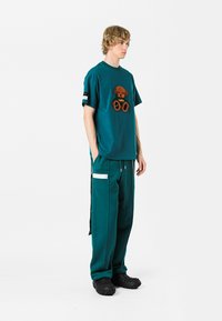 Camiseta de color verde azulado con un gráfico texturizado marrón y detalles en negro, combinada con pantalones jogger a juego en verde azulado con bolsillos con cremallera y detalles laterales.
