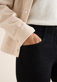 Ljusbeige corduroyjacka med ribbad textur, knäppta ärmslut och en vit stickad tröja under, tillsammans med åtsittande svarta jeans.
