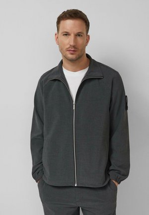 Man draagt een donkergrijs jack met ritssluiting over een wit shirt, met handen in de zakken, staand tegen een effen lichtgrijze achtergrond.