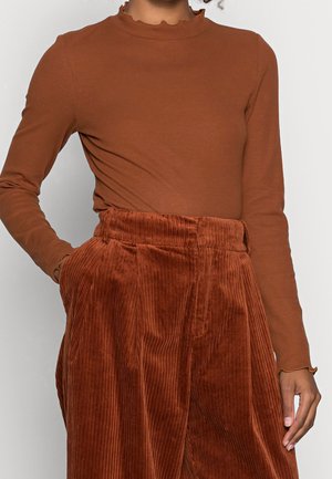 Femme portant un haut côtelé à manches longues couleur rouille avec un col à volants et un pantalon en velours côtelé rouille taille haute avec des poches.