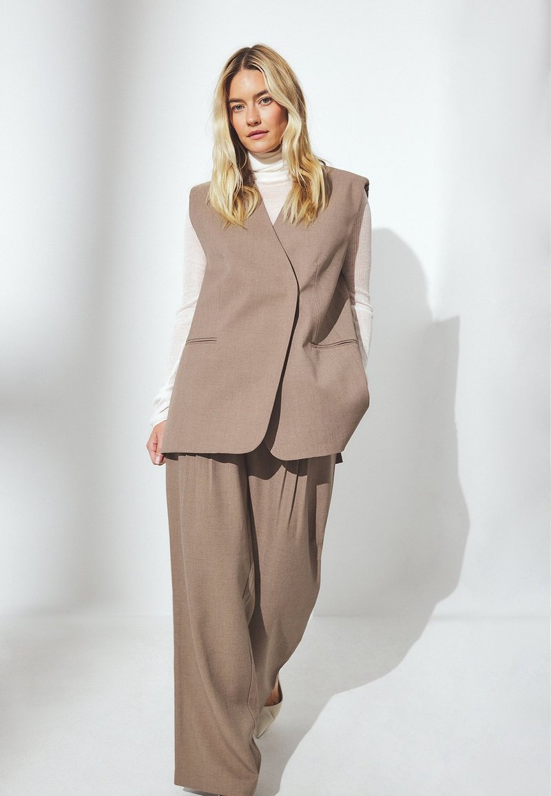 Beige tailored vest with a structured design, featuring a v-neck, side pockets, and wide-leg trousers, all in a soft, textured fabric.