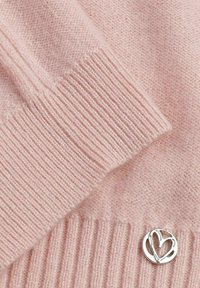 Pull en cachemire rose clair avec des poignets côtelés. Présente un charm en forme de cœur argenté sur l'ourlet. Texture douce et tricot délicat.