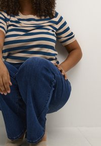 Stribet kortærmet sweater i beige og marineblå, parret med blå denim jeans. Nærbillede af en person, der sidder med krydsede ben og hænderne på knæene.