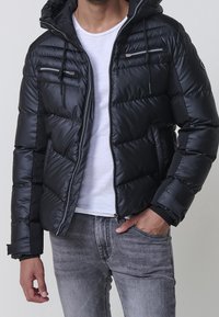 Homme portant une veste matelassée noire à capuche avec poches zippées, t-shirt blanc et jean gris, main dans la poche, sur fond uni.