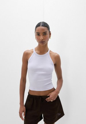 Femme aux cheveux foncés portant un haut côtelé blanc sans manches et un short noir à cordon se tient devant un fond clair uni.