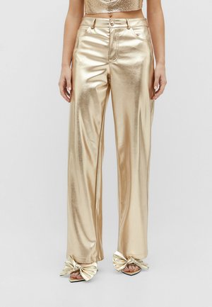 Vrouw draagt glanzende gouden broek met wijde pijpen, gecombineerd met bijpassende gouden sandalen met grote strikaccenten.