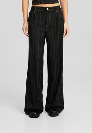 Sorte bukser med vide ben og tynde hvide lodrette nålestriber, båret med sorte sko, vist fra talje til fødder på en neutral baggrund.