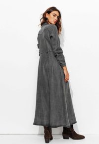 Robe en denim gris longue avec des manches longues, un col à revers et une taille cintrée. Se termine aux chevilles, avec un ourlet effiloché et des fentes latérales.