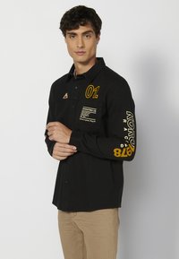 Camisa negra de manga larga con gráficos amarillos, un bolsillo de parche con texto y un cuello puntiagudo. La tela es suave y con un corte estructurado.