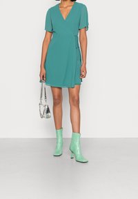Robe portefeuille en bleu canard avec des manches courtes à volants, associée à des bottines vertes à talons hauts et un sac à main argenté. Tissu lisse et coupe structurée.