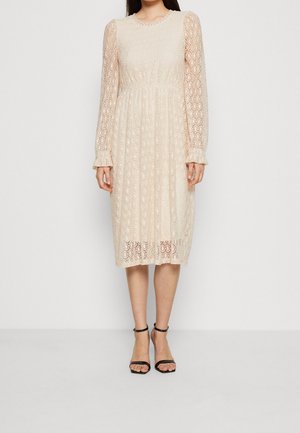 Spitzen-Midikleid in hellem Beige mit langen Ärmeln, rundem Ausschnitt, filigranen Mustern und ausgestelltem Rock. Schwarze Riemchensandalen darunter getragen.