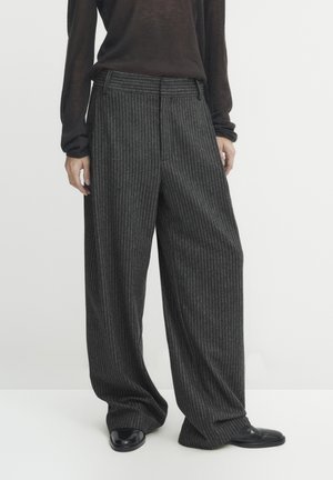 Person iført løstsiddende mørkegrå bukser med lyse striber og vide ben samt sorte sko, stående mod en ensartet lys baggrund.