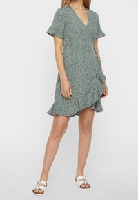 Zelené šaty s překříženým designem, krátkými rukávy a volánkovým lemem; mají rozptýlený vzor bílých teček a jsou vyrobeny z lehkého materiálu.