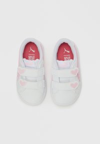 Λευκά παιδικά αθλητικά παπούτσια με ροζ αυτοκόλλητα λουριά σε σχήμα καρδιάς και κόκκινες εσώσολες που φέρουν το λογότυπο της Puma και QR κώδικα,  απεικονιζόμενα από πάνω.