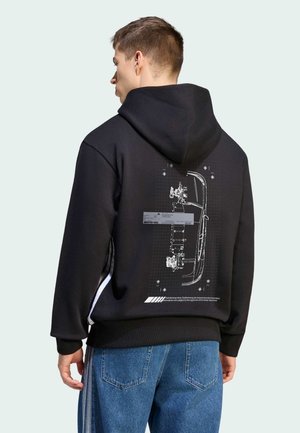 Mand iført en sort hættetrøje med teknisk biltegning på ryggen, kombineret med blå jeans mod en ensfarvet baggrund.