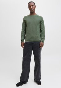 Man standing wearing olive green long-sleeve sweater, loose-fitting black jeans, and black shoes against plain light background.