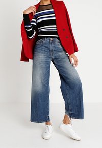 Weite blaue Jeans aus Denim mit fransigem Saum, kombiniert mit einem schwarzen, gestreiften, taillierten Oberteil und einem roten Blazer. Weiße Sneakers runden das Outfit ab.