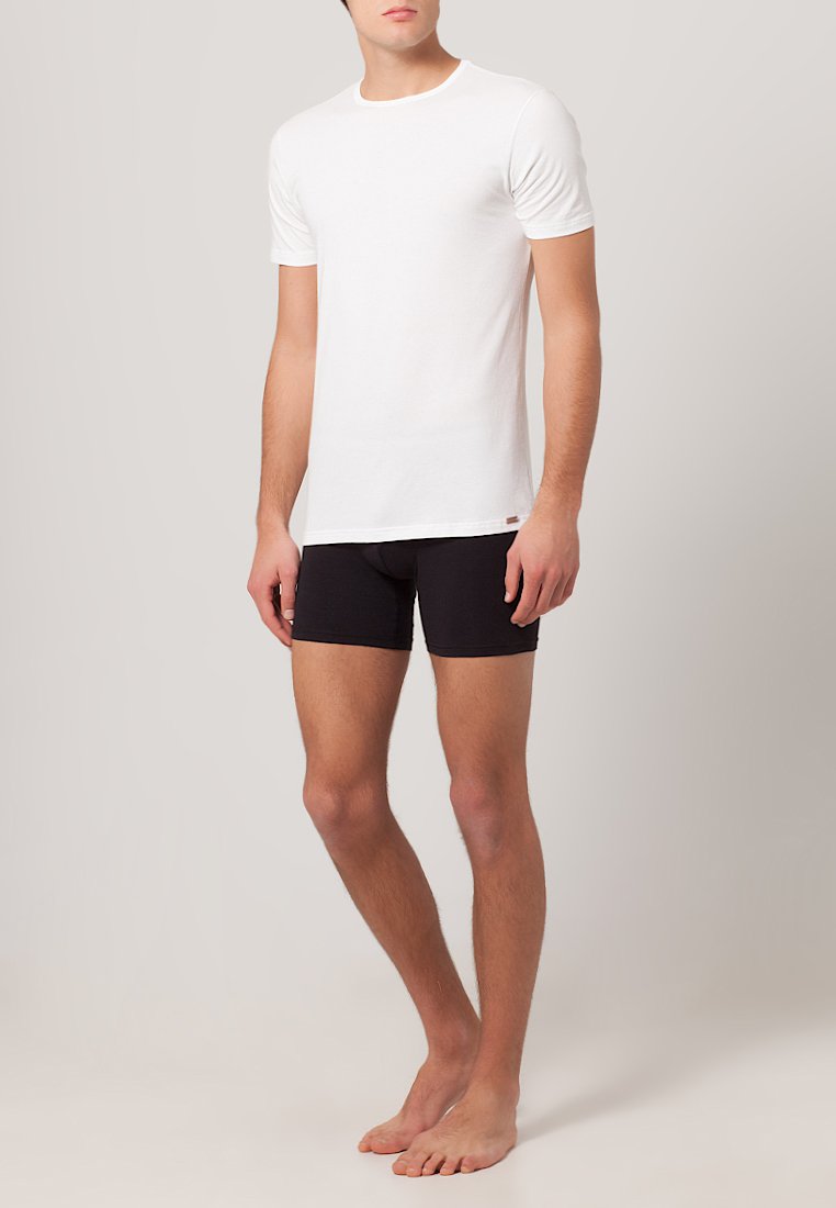T-shirt bianca in cotone con maniche corte e scollatura a girocollo, abbinata a pantaloni boxer neri. Il modello è in piedi a piedi nudi su uno sfondo neutro.