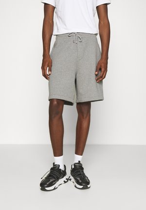 Mężczyzna ubrany w jasno szare szorty na sznurku, białe skarpetki, czarno-białe trampki oraz białą koszulkę, stojący na jednolitym tle.