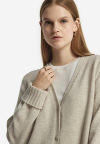 Beige kardigan med räfflade manschetter, mjuk textur och V-ringad design, tillsammans med en ljus T-shirt under.