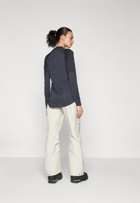 Chemise à manches longues bleu marine avec des accents bleu clair, assortie à un pantalon beige avec des zippers, portée avec des bottes noires.
