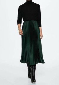 Black turtleneck paired with a knee-length, pleated green skirt, featuring a smooth texture and a flowy silhouette. Black knee-high boots complete the look.