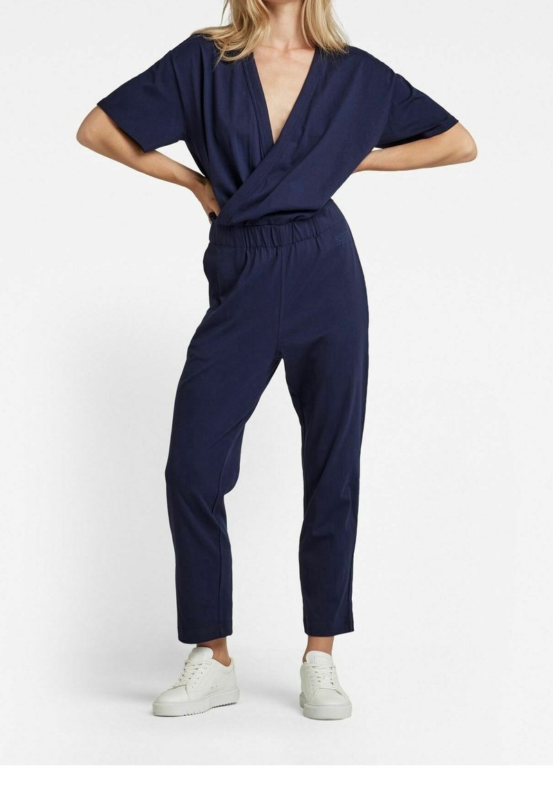 Femme en combinaison bleu marine style portefeuille à manches courtes avec des baskets blanches, debout les mains sur les hanches sur fond blanc.