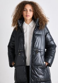 Casaco acolchoado em material preto brilhante, apresenta uma gola alta, fecho de correr e dois bolsos frontais com detalhes em dourado.