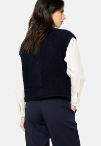 Jūras zila, pūkaina veste ar biezu, teksturētu audumu; bez piedurknēm, nēsāta virs baltas garas piedurknes krekla, kombinēta ar tumšiem biksēm.