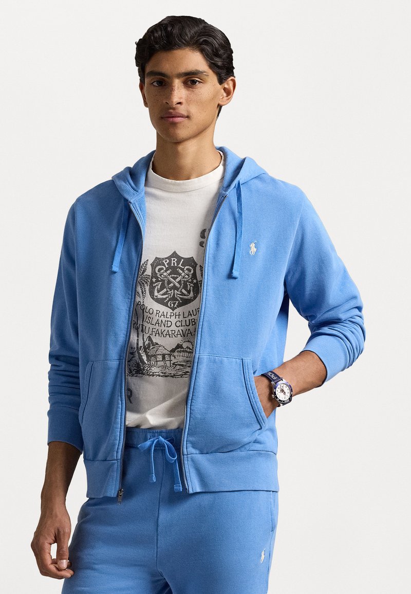 Ανοιχτό μπλε αθλητική ζακέτα με φερμουάρ από μαλακό ύφασμα, φορεμένη πάνω από ένα λευκό μπλουζάκι με γραφικά. Συνοδεύεται από ταιριαστά φόρμα και ρολόι.