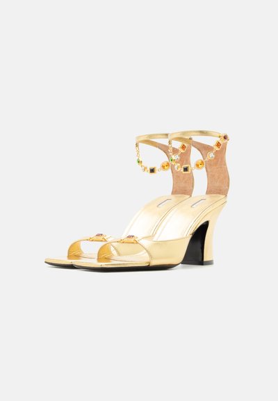 Zlaté sandále na vysokom opätku s otvorenou špičkou a klenotmi zdobenými remienkami na členky s viacfarebnými drahokamami a lesklým povrchom.