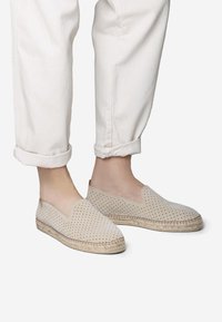 Beige geperforeerde instapschoenen met jutezool; afgeronde voorkant, geen zichtbare hardware en bovenwerk van textiel, gecombineerd met opgerolde witte denimbroeken.